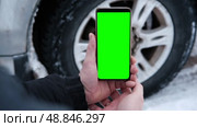 Male hands holding a smartphone with a bright green screen against the background of a car wheel. Automotive technology. Mobile applications for drivers. Road safety. Using gadgets. Chroma key. Стоковое видео, видеограф Евгений Демидюк / Фотобанк Лори