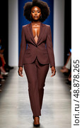 Confident model in a pinstripe suit walks down a fashion runway, flaunting bold afro hairstyle. Audience members in the background with blurred details enhance focus on the model. Стоковое AI-изображение, AI-художник Olga Dmitrieva / Фотобанк Лори