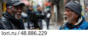 A close-up shot of two men, both with gray beards and wearing hats, engaged in filming on a city street. Стоковое AI-изображение, AI-художник Данила Большаков / Фотобанк Лори