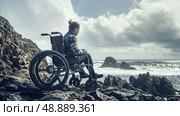 A woman in a wheelchair sits on a rocky coastline overlooking the ocean. The cloudy sky and turbulent water create a dramatic backdrop for her contemplative pose. Стоковое AI-изображение, AI-художник Данила Большаков / Фотобанк Лори