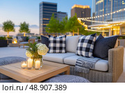Cozy outdoor lounge area featuring stylish seating with cushions, decorative poufs, and ambient lighting, surrounded by greenery and modern architecture, perfect for relaxation and social gatherings. Стоковое AI-изображение, AI-художник Ирина Ручушкина / Фотобанк Лори