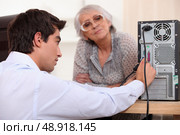 young man wearing a shirt is plugging cables on the back of a computer, an old woman is watching him. Стоковое фото, агентство Ingram Publishing / Фотобанк Лори