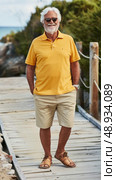 A senior man with a white beard is smiling and walking on a wooden path wearing a yellow polo shirt, khaki shorts, sunglasses, and sandals. Стоковое AI-изображение, AI-художник Данила Большаков / Фотобанк Лори