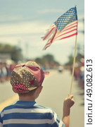 A young boy in a striped shirt and patterned hat holds an American flag high during a parade. The flag waves in the wind. Стоковое AI-изображение, AI-художник Данила Большаков / Фотобанк Лори