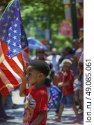 Young boy salutes an American flag during a public celebration. He is wearing a red shirt and standing in a crowd of people. The scene is a festive event showcasing patriotism. Стоковое AI-изображение, AI-художник Данила Большаков / Фотобанк Лори