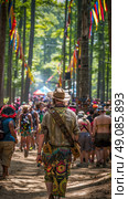 A man wearing a hat and colorful pants walks through a wooded festival area with colorful flags hanging in the trees. Стоковое AI-изображение, AI-художник Данила Большаков / Фотобанк Лори