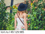 A young woman wearing a hat and a dress is admiring some sunflowers outside a house on a sunny day in summer. Стоковое фото, агентство Ingram Publishing / Фотобанк Лори