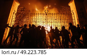 Silhouetted figures gather by a blazing fire during a nighttime protest outside grand gates. Стоковое AI-изображение, AI-художник Данила Большаков / Фотобанк Лори