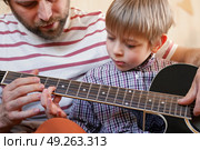 Handsome dad teaches his cute son to play the guitar sitting on sofa in the living room at home. Happy Father's Day. Father teaching his child to play guitar. Kid Learning music skill. Art education. Стоковое фото, фотограф Марина Демидюк / Фотобанк Лори