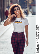 Smiling young arabic woman with black curly hairstyle. Arab girl in casual clothes in the street. Happy female wearing white t-shirt and checked pants. Стоковое фото, агентство Ingram Publishing / Фотобанк Лори
