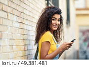 Young Arab woman texting with her smart phone on brick urban wall. Smiling North Afrincan girl in casual clothes with black curly hairstyle. Стоковое фото, агентство Ingram Publishing / Фотобанк Лори