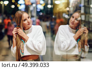 Blonde girl wearing white sweater standing in the street with defocused city lights at the background. Pretty woman with pigtail hairstyle at night reflected in a shop window. Стоковое фото, агентство Ingram Publishing / Фотобанк Лори