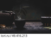 Dark tea background with black iron asian teapot and mug of hot tea with steam on table. Copy space for your design. Authentic vintage style. Traditional tea ceremony arrangement. Стоковое фото, агентство Ingram Publishing / Фотобанк Лори