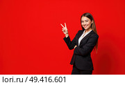 Улыбающаяся девушка показывает два пальца. Красивая молодая женщина в деловом костюме на ярком красном фоне. Стоковое AI-изображение, AI-художник Илья Старков / Фотобанк Лори