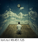 Abstract idea with a person sitting in a dark room in front of a clock surrounded by limitations daily routine concrete walls with clouds texture. Time pressure deadline concept. Стоковое фото, агентство Ingram Publishing / Фотобанк Лори