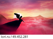 Silhouette of a lonely fallen angel with long wings standing on a cliff against a paradise sunset. Dusk sky over the clouds in the mountains. Heaven landscape scene screen saver. Стоковое фото, агентство Ingram Publishing / Фотобанк Лори