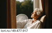 Senior woman enjoying warm sunlight by the window, eyes closed, wearing a white shirt, captured in a peaceful and serene moment of relaxation indoors. Стоковое AI-изображение, AI-художник Катерина Бондарец / Фотобанк Лори
