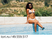 Relaxed woman sunbathing sitting on the edge of a swimming pool with sunglasses, tanned body, curly long hair, fashionable bikini. Стоковое фото, агентство Ingram Publishing / Фотобанк Лори