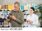 Senior man in pharmacy scans QR code on antitussive syrup, girl pharmacist in background. Стоковое фото, фотограф Яков Филимонов / Фотобанк Лори