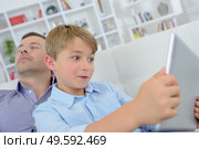 Child wide eyed looking at tablet, father asleep. Стоковое фото, агентство Ingram Publishing / Фотобанк Лори
