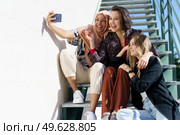 Trendy young cheerful Muslim lady in stylish outfit and traditional hijab, smiling brightly while taking selfie on mobile phone, sitting on staircase with diverse girlfriends on sunny day. Happy multiethnic female best friends taking selfie sitting on stairs in. Стоковое фото, агентство Ingram Publishing / Фотобанк Лори