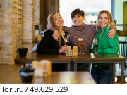 Cheerful adult female with short dark hair looking at camera with smile and embracing girlfriends behind table with alcohol during meeting in pub. Happy women hugging during party in bar. Стоковое фото, агентство Ingram Publishing / Фотобанк Лори