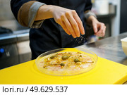 Soft focus of anonymous cook scattering salt over plate of prawn carpaccio with capers during work in restaurant kitchen. Crop chef garnishing dish of prawn carpaccio with capers. Стоковое фото, агентство Ingram Publishing / Фотобанк Лори