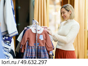 Side view of smiling female customer choosing apparel for girl while standing near rack with various stylish clothes in modern boutique. Content woman choosing clothes for little girl. Стоковое фото, агентство Ingram Publishing / Фотобанк Лори