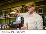 Low angle of bearded man in white shirt and cap filling glass goblet with fruit cocktail while working in bar in evening. Barman pouring cocktail into glass. Стоковое фото, агентство Ingram Publishing / Фотобанк Лори