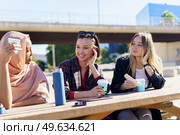 Smiling young multiethnic female students in casual clothes chating in an outdoor cafe and drinking coffee from take-away cups. Smiling diverse young women having coffee break and chatting in city park. Стоковое фото, агентство Ingram Publishing / Фотобанк Лори