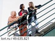 Low angle of trendy young multiethnic women best friends, on stylish outfit and hijab, leaning on metal railing while standing on staircase and looking away in city on sunny day. Content young diverse women looking away while standing on stairs in city. Стоковое фото, агентство Ingram Publishing / Фотобанк Лори
