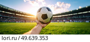 A hand holding a soccer ball in a stadium at sunset symbolizing passion for sports, global football culture, and the spirit of the game. Стоковое AI-изображение, AI-художник Igor Tichonow / Фотобанк Лори