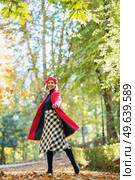 Full body of active female in red coat and beret looking at camera while standing on pathway on autumn day. Smiling woman in autumn park. Стоковое фото, агентство Ingram Publishing / Фотобанк Лори