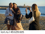 Anonymous woman taking photo of positive multiracial female friends hugging while standing on sandy coast near rippling sea on sunny day. Faceless woman photographing cheerful diverse girlfriends on seashore. Стоковое фото, агентство Ingram Publishing / Фотобанк Лори