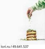 Woman hand sprinkles fresh cress sprouts above layered club sandwich at white background. Preparing healthy lunch.  Front view. Стоковое фото, агентство Ingram Publishing / Фотобанк Лори