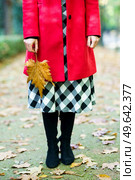 Crop anonymous female wearing red coat and checkered skirt standing with yellow leaf on pathway in autumn park. Woman with dry leaf standing on pathway. Стоковое фото, агентство Ingram Publishing / Фотобанк Лори