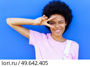 Positive African American female with Afro hairstyle showing V sign near eyes while winking and looking away against blue background. Cheerful black woman making peace sign near wall. Стоковое фото, агентство Ingram Publishing / Фотобанк Лори