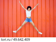 Full body of delighted African American female in casual wear looking at camera while jumping with raised arms against red background. Happy black woman jumping on street. Стоковое фото, агентство Ingram Publishing / Фотобанк Лори