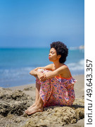 Tranquil ethnic female in maxi dress with curly hair and closed eyes, embracing knees while sitting on stones against waving sea and blue sky on summer weekend day on beach. Calm black woman resting against sea. Стоковое фото, агентство Ingram Publishing / Фотобанк Лори