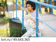 Side view of wistful African American female in casual outfit looking into distance with thoughtful face while sitting on footbridge against blurred background. Dreamy black woman sitting on footbridge. Стоковое фото, агентство Ingram Publishing / Фотобанк Лори