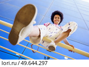 From below full body of positive African American female in overall looking at camera while sitting on fence near glass building. Smiling black woman sitting on railing. Стоковое фото, агентство Ingram Publishing / Фотобанк Лори