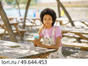 Wistful African American female with takeaway coffee looking away while sitting at wooden table in park on sunny summer day. Black woman with coffee in park. Стоковое фото, агентство Ingram Publishing / Фотобанк Лори