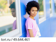Serious African American female with Afro hairstyle looking away with thoughtful face while standing near blue building with windows on street of city. Black woman standing near blue house. Стоковое фото, агентство Ingram Publishing / Фотобанк Лори