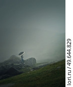 Man with umbrella climbing a mountain hill in a gloomy weather with dense mist. Moody hiking scene with a wanderer walking on the cliffs of the foggy valley. Стоковое фото, агентство Ingram Publishing / Фотобанк Лори