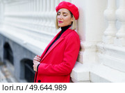 Dreamy female with blond hair in red clothes with beret standing with closed eyes near white fence on street of city. Charming woman in red outfit near fence. Стоковое фото, агентство Ingram Publishing / Фотобанк Лори
