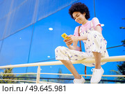 Glad African American female with Afro hairstyle in overall browsing cellphone while sitting on railing near glass building in city. Content black woman texting message on fence. Стоковое фото, агентство Ingram Publishing / Фотобанк Лори