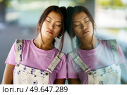 Calm stylish Asian female closing eyes and leaning on reflective glass wall on sunny day on city street. Asian woman near glass wall. Стоковое фото, агентство Ingram Publishing / Фотобанк Лори