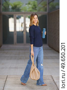Full body positive stylish woman with thermos and bag closing eyes and smiling while walking on pavement outside modern building. Stylish female smiling and walking on pavement. Стоковое фото, агентство Ingram Publishing / Фотобанк Лори