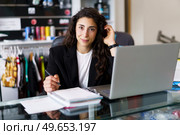 Positive brunette looking at camera and adjusting long wavy hair while sitting at desk with netbook and making notes during psychology session in gym. Glad sports psychologist touching hair during work. Стоковое фото, агентство Ingram Publishing / Фотобанк Лори