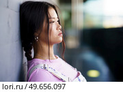 Young Asian female in casual clothes with braid leaning on wall and looking away on city street in daytime. Asian woman leaning on wall. Стоковое фото, агентство Ingram Publishing / Фотобанк Лори